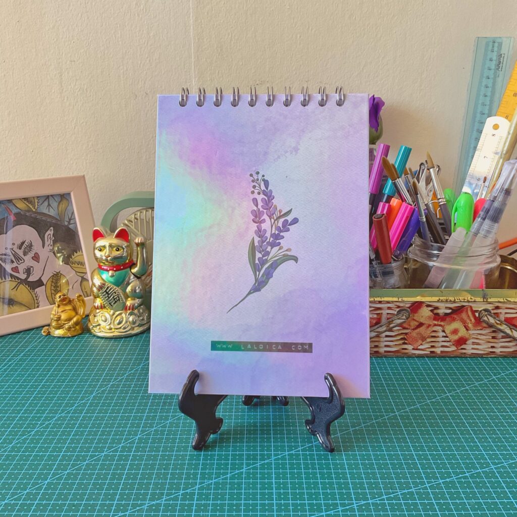 Libreta anillada vertical con ilustración de tapa trasera de lavanda sobre fondo acuarela en tonos lilas, celestes y rosados. Está sobre un atril negro, con un escritorio de fondo decorado con lápices, reglas, figuras de la suerte y una ilustración enmarcada.