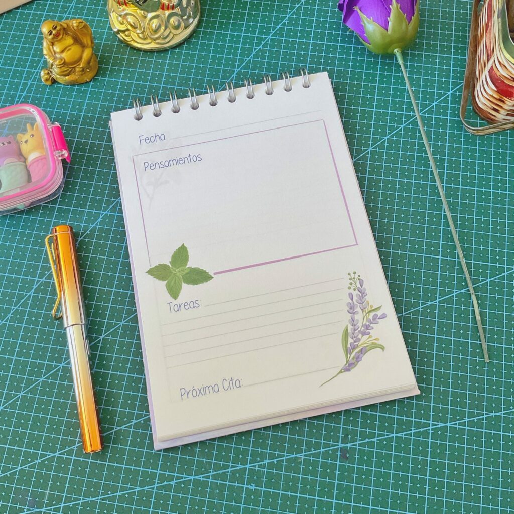 Página interior del cuaderno de terapias con secciones tituladas “Fecha”, “Pensamientos”, “Tareas” y “Próxima Cita”, decorada con ilustraciones de lavanda y hojas verdes. Está sobre una base de corte verde con objetos como un lapiz naranja y figuras decorativas alrededor.