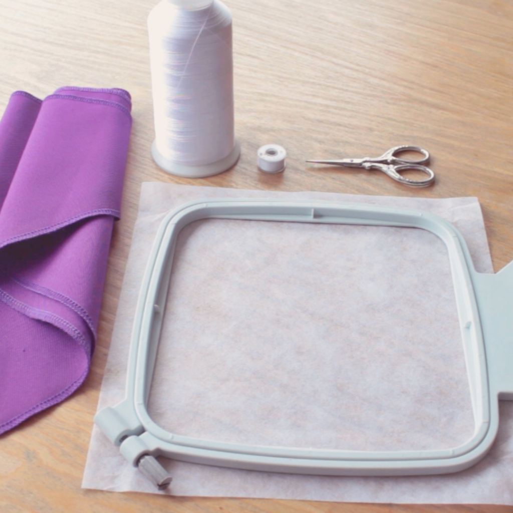 Materiales para bordar: un pañuelo morado doblado, un carrete de hilo blanco, una bobina blanca, tijeras pequeñas y un bastidor de bordado con estabilizador colocado, todos sobre una superficie de madera.