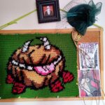 Fotografiá de pizarra de corcho colgada en una pared blanca, sobre la pizarra se encuentra colgado un tejido a crochet/ganchillo del personaje Chester